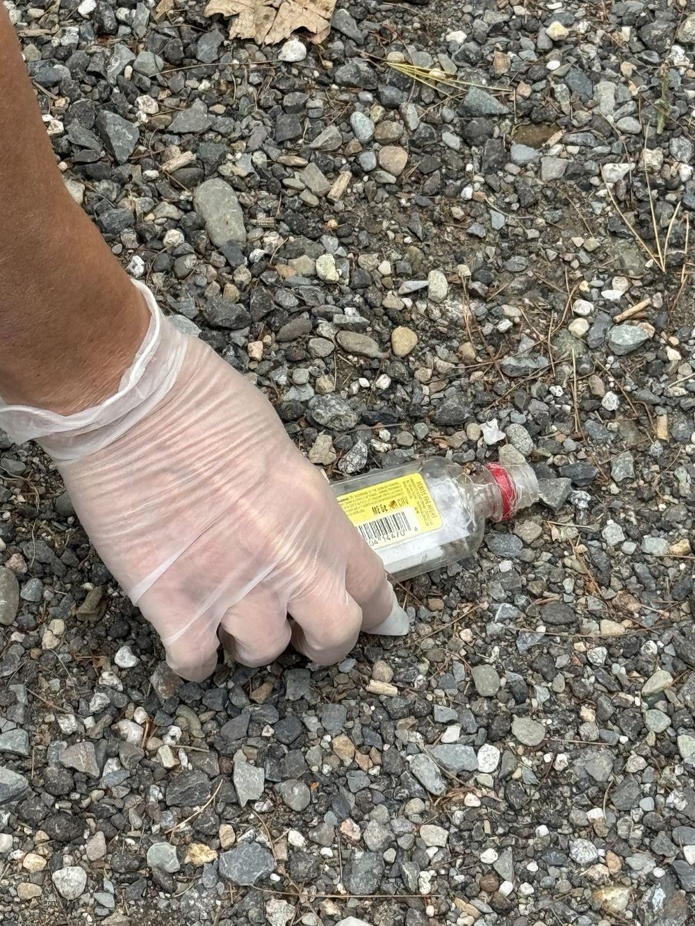 A gloved hand picking up a small empty bottle from a gravel shoulder. The label shows the Maine 5 cent deposit mark.